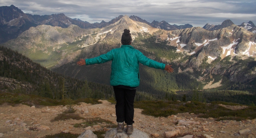 A person facing away from the camera spreads their arms out wide while standing on a rock overlooking a vast mountainous landscape.